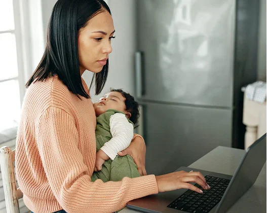 A woman working on a laptop while holding a sleeping baby